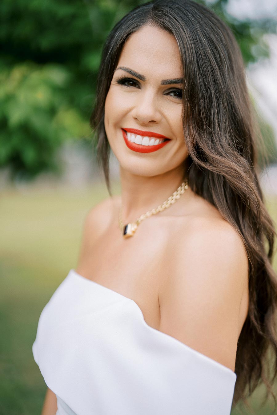 Smiling woman with long brown hair wearing a white off-shoulder dress and a gold necklace, standing outdoors with a blurred green background.