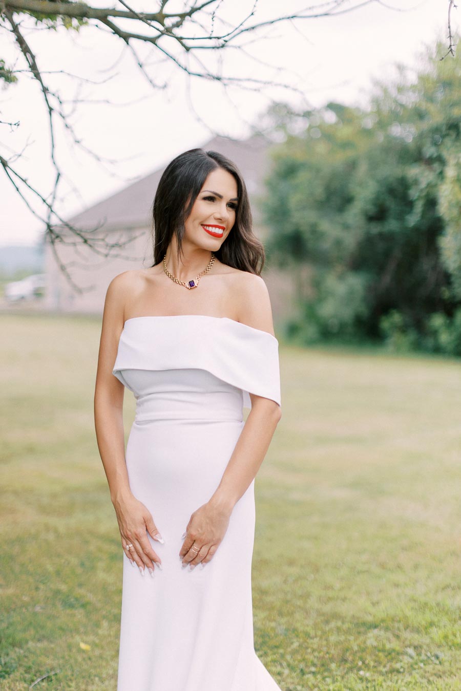 Elegant woman in a white off-shoulder dress smiling outdoors, wearing a necklace, with a blurred green background.