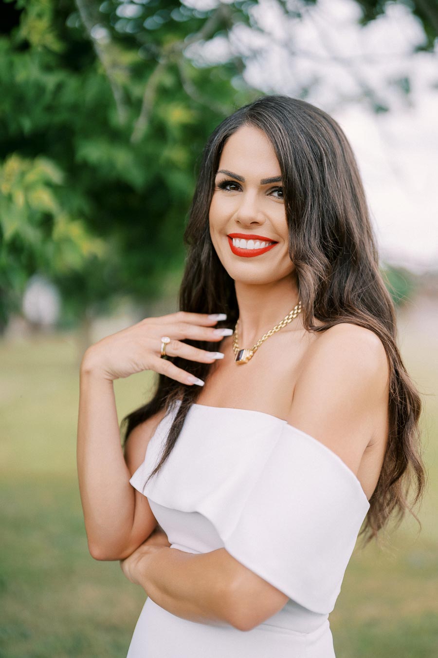 Smiling woman with long dark hair wearing a white off-the-shoulder dress and red lipstick, standing outdoors with a green leafy background.
