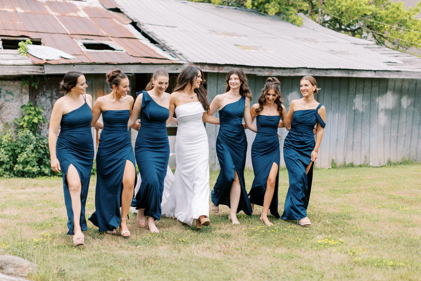 Bride in a white gown with bridesmaids in blue dresses walking on a grassy area, rustic barn in the background.
