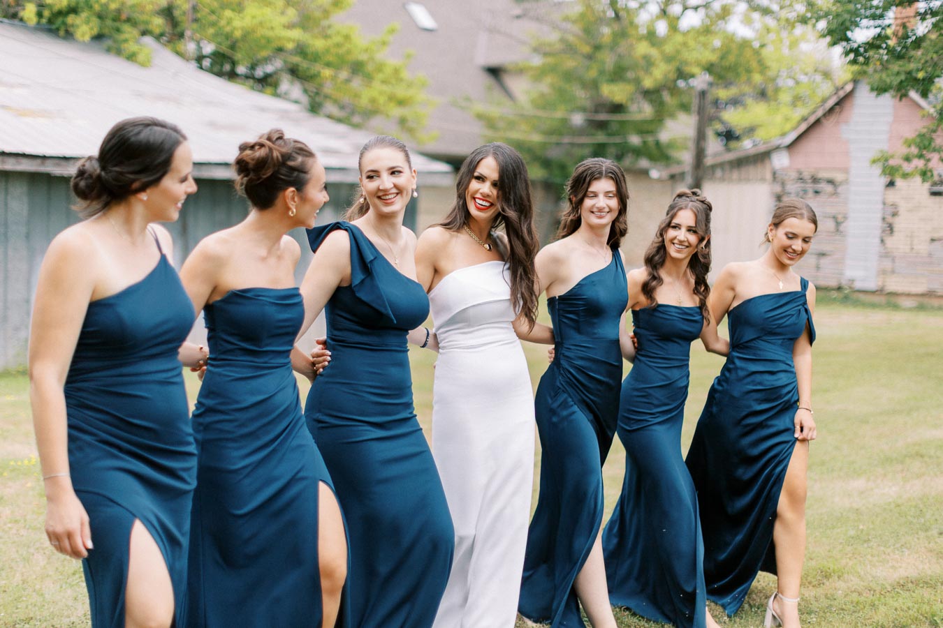 A bride in a white dress happily walks with her bridesmaids in matching navy blue dresses with one-shoulder designs, outdoors on a grassy area with rustic buildings in the background.