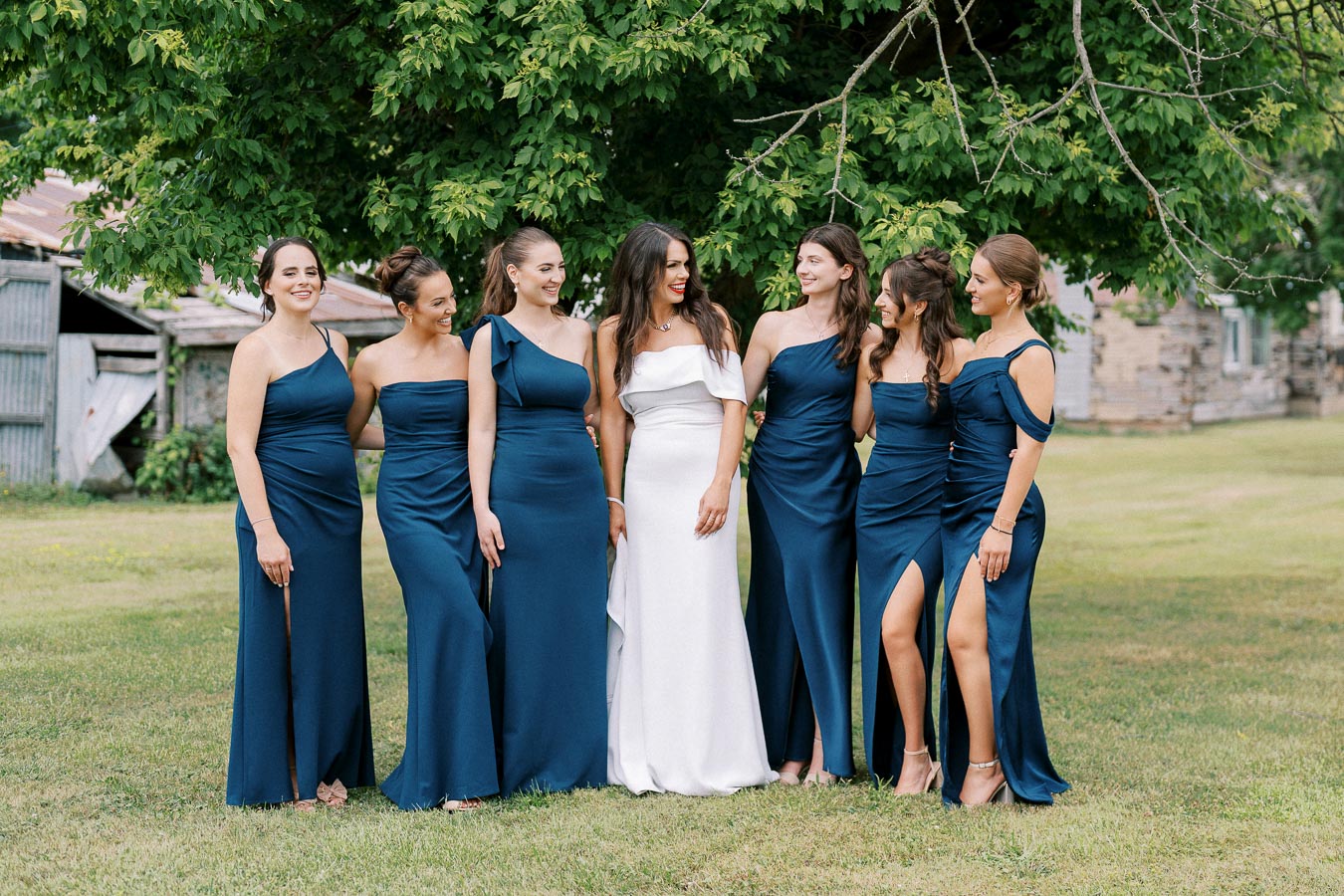Group of bridesmaids in matching navy blue dresses with a bride in a white gown standing together outdoors, smiling under a large tree with a rustic barn in the background.