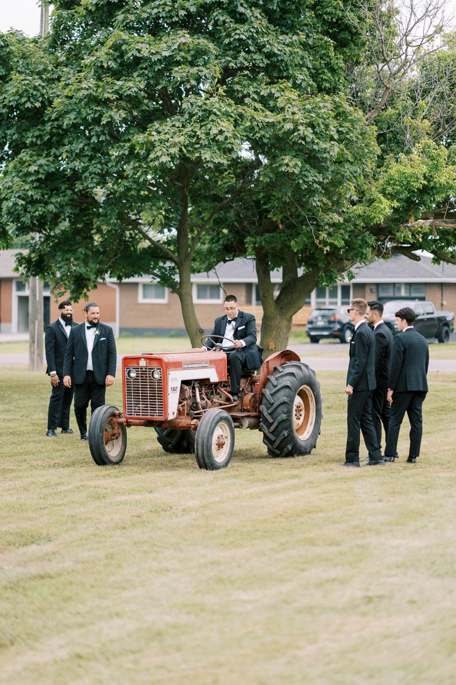 A group of men in formal suits gathered around a vintage red tractor on a sunny day, with a lush green tree and suburban houses in the background.