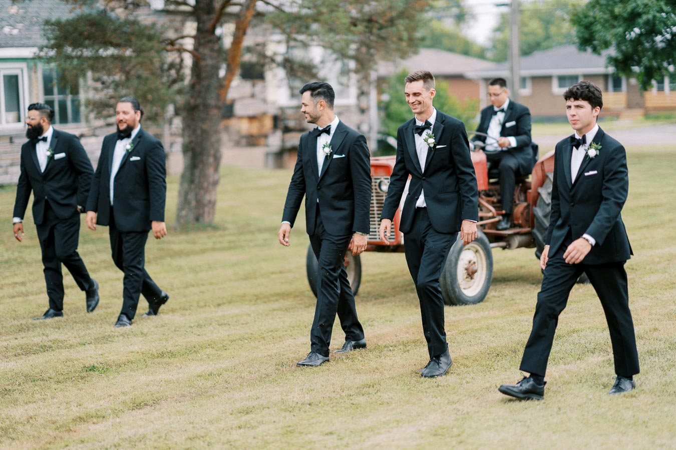 Groomsmen in black tuxedos walking across a grassy field with a vintage tractor in the background, at an outdoor wedding.