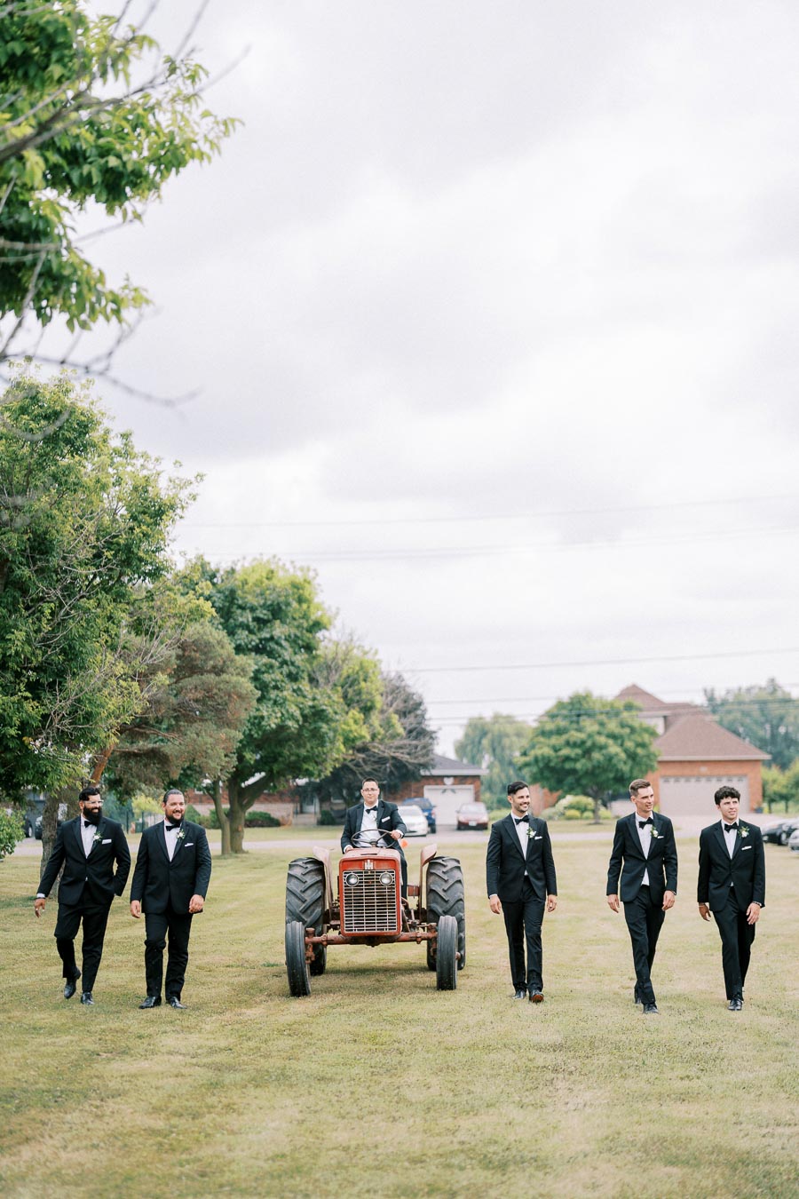 Groomsmen in tuxedos walking with a vintage tractor in a grassy outdoor setting, surrounded by trees and houses under a cloudy sky.