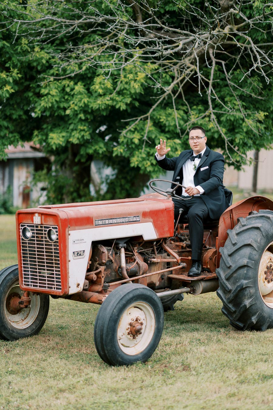 A person in a tuxedo sits on an old red tractor waving, surrounded by lush green trees in a rustic outdoor setting.