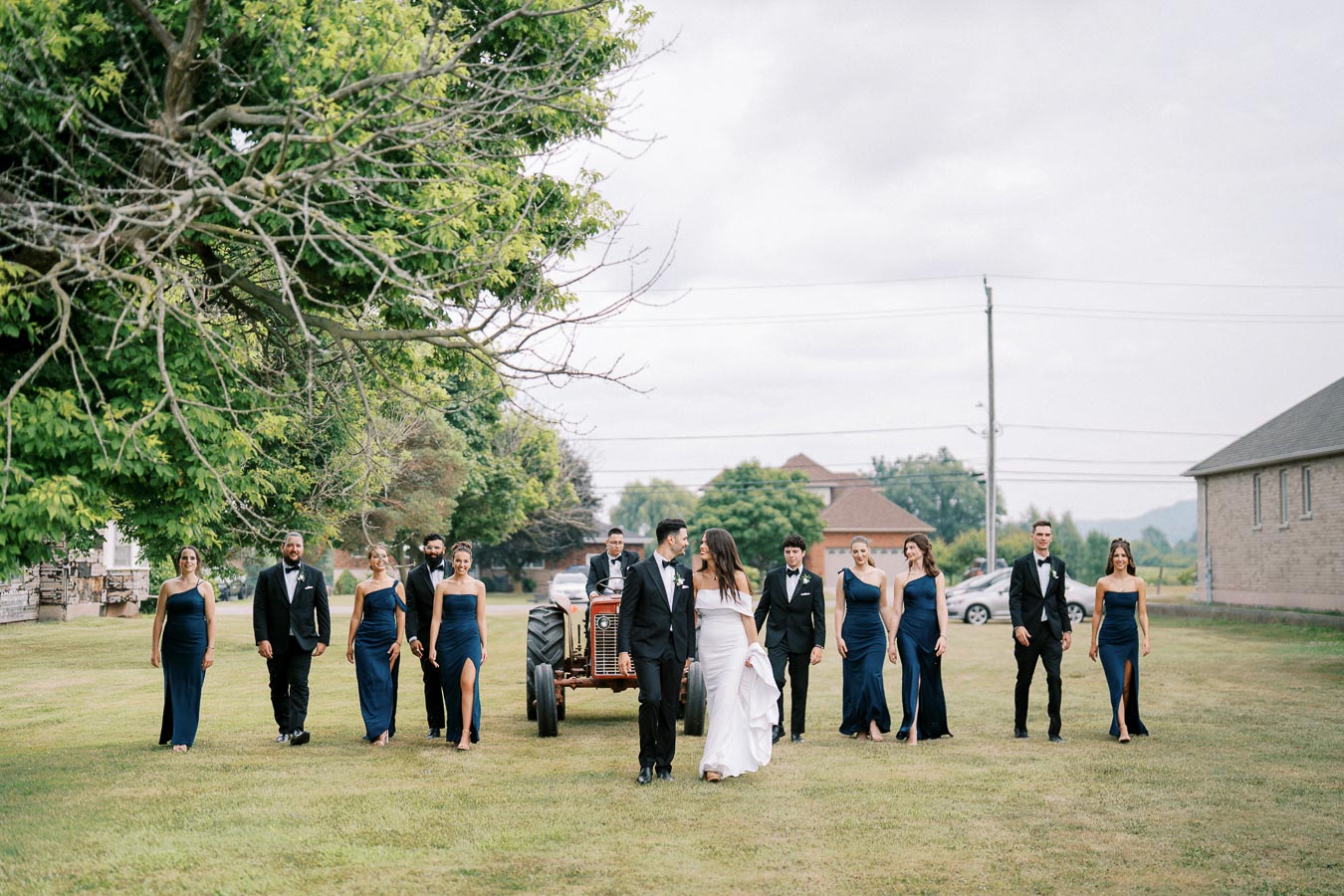 Wedding party walking outdoors on a grassy lawn, featuring bridesmaids in matching navy blue dresses and groomsmen in black tuxedos on a cloudy day, with lush green trees in the background.