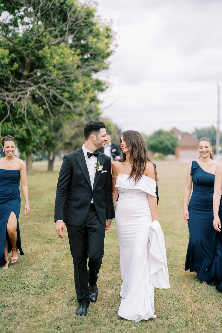 A bride in a white gown and groom in a black tuxedo walk together on a lawn, smiling at each other, with bridesmaids in navy dresses following behind under a cloudy sky.