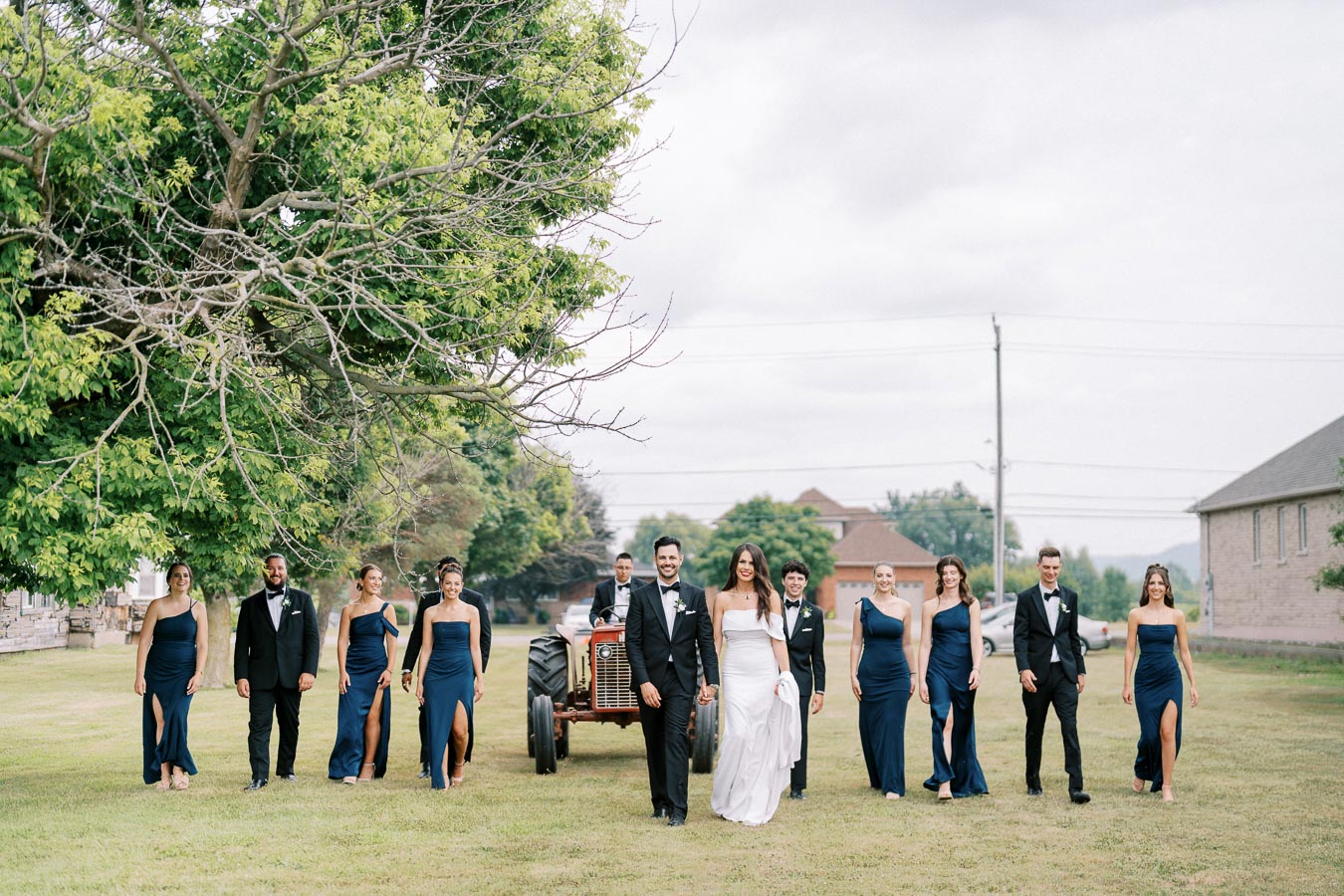 Wedding party walking outdoors on a grassy field, featuring a bride in a white dress and groom in a black tuxedo, accompanied by bridesmaids in navy dresses and groomsmen in black suits. A rustic tractor and lush green trees are visible in the background, creating a picturesque countryside wedding setting.