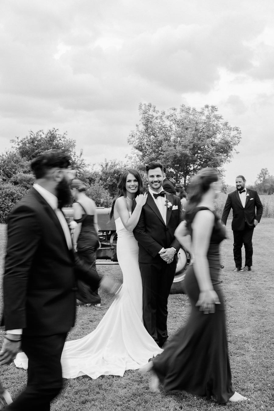 A bride in a white dress and groom in a suit stand together smiling outdoors on their wedding day, surrounded by guests in formal attire, captured in a candid black and white photograph.