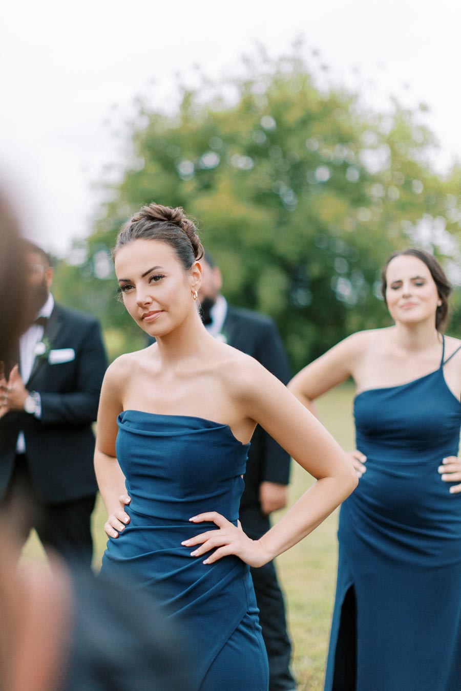 Young woman in a blue strapless dress standing outdoors with hands on hips, surrounded by people in formal attire, and blurred greenery in the background.