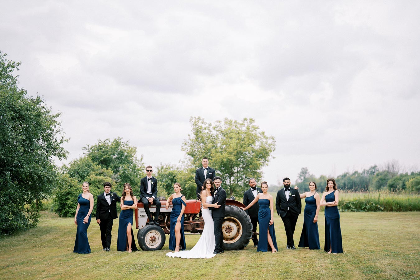 Wedding party posing outdoors with a vintage tractor in a lush green field, bridesmaids in elegant blue dresses, and groomsmen in tuxedos.