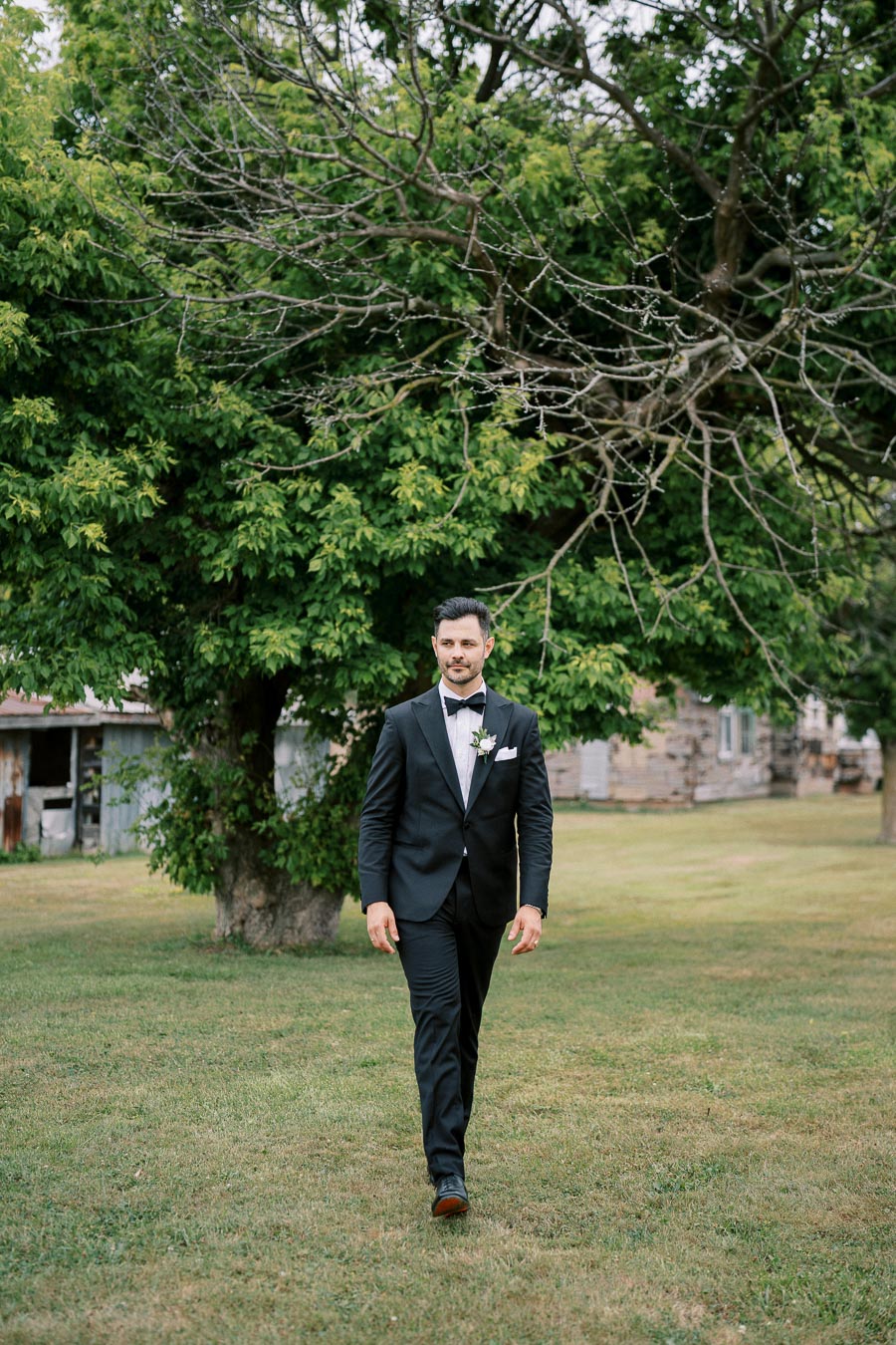 A man in a formal black suit with a white shirt and bow tie walking on a grassy lawn, framed by lush green trees and rustic buildings in the background.
