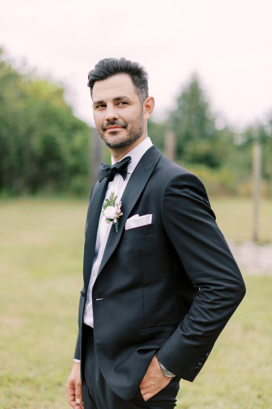 Man in a black tuxedo with bow tie standing outdoors, wearing a white dress shirt and floral boutonniere, surrounded by greenery background.