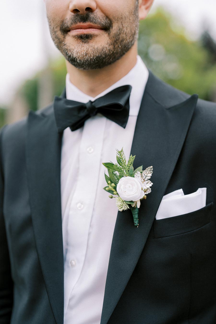 A man wearing a formal black tuxedo with a white dress shirt, black bow tie, and a white rose boutonniere, ideal for a wedding or formal event.
