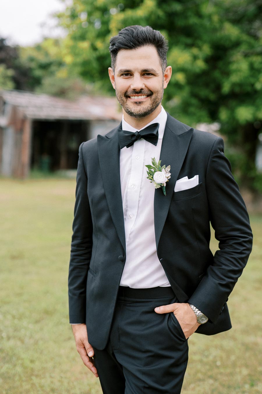A groom in a sleek black tuxedo with a white shirt and bow tie, smiling outdoors with greenery in the background, showcasing a white boutonniere on his lapel, embodying elegance and style for a wedding day.