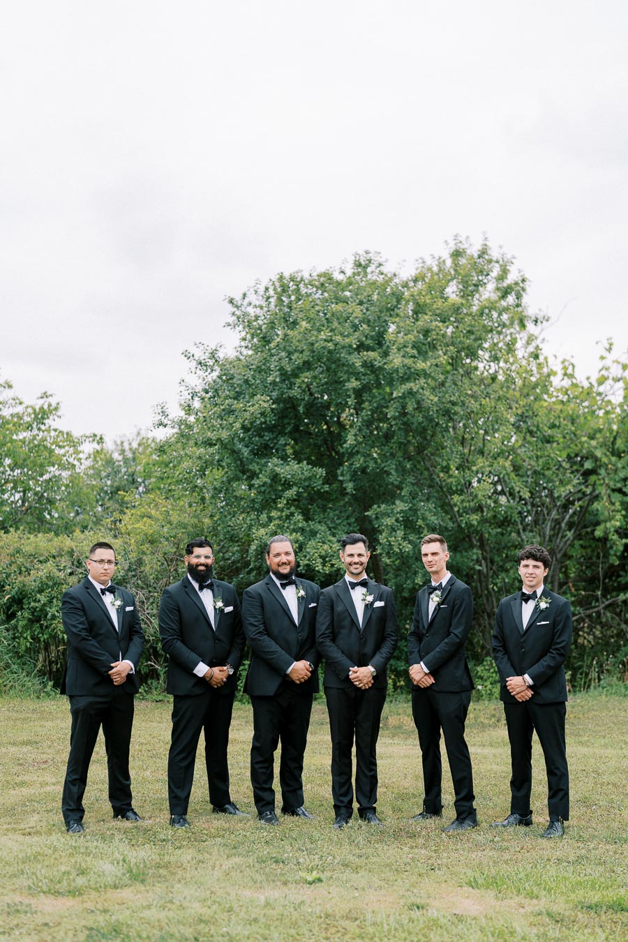 Groomsmen standing in a line outdoors, wearing black tuxedos and bow ties, with greenery and trees in the background.