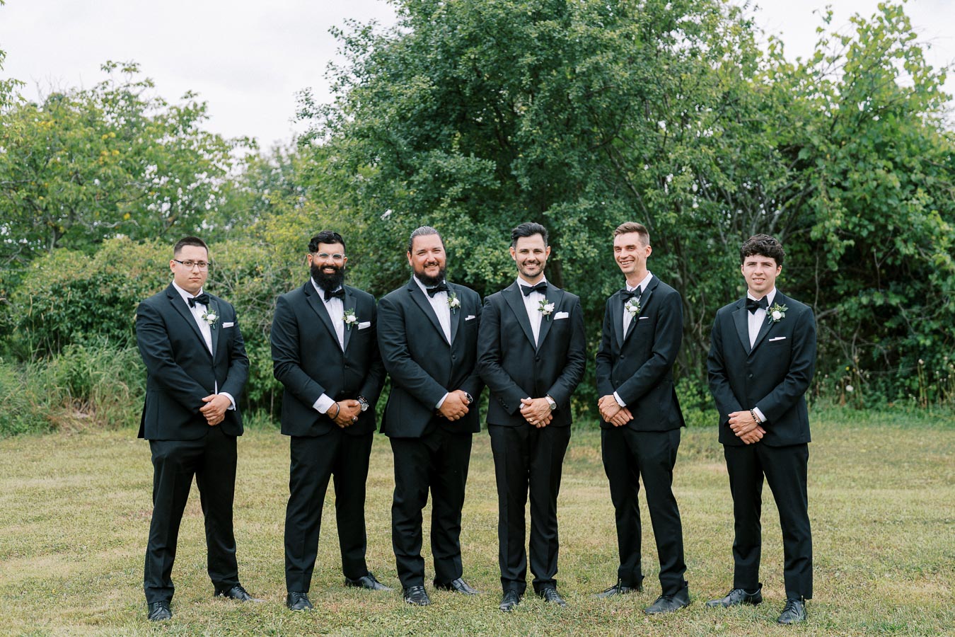 A group of six groomsmen in black tuxedos and bow ties standing together on a grassy lawn with green trees in the background, showcasing a formal outdoor wedding setting.