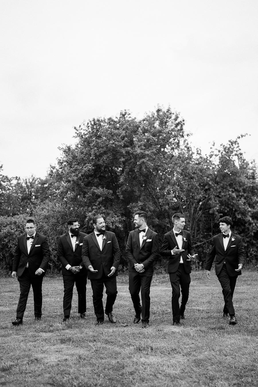 Groomsmen in black tuxedos walking together on a grassy field, set against a backdrop of trees.