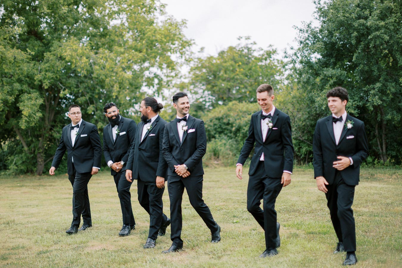 A group of six groomsmen walking together outdoors in a grassy area, wearing matching black suits and bow ties, surrounded by lush green trees in the background.