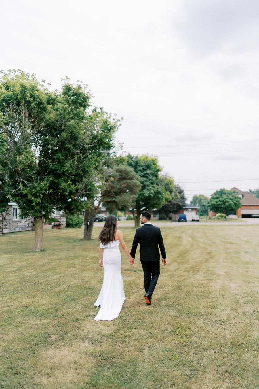 Wedding couple walking hand in hand on a grassy field, bride in white dress and groom in black suit, surrounded by trees on a cloudy day.
