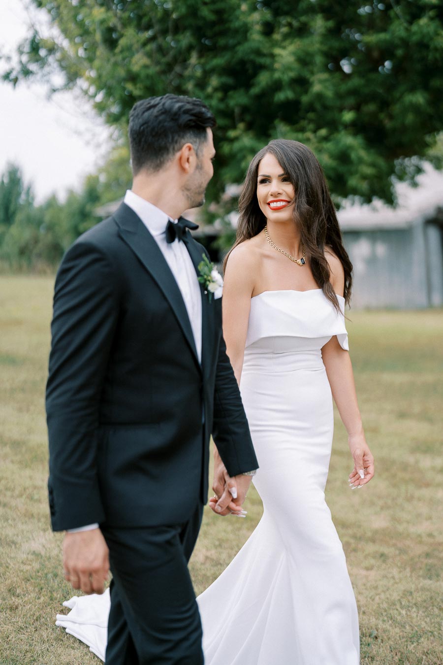 A bride in a white dress and a groom in a black tuxedo smiling at each other while holding hands outdoors with greenery in the background.