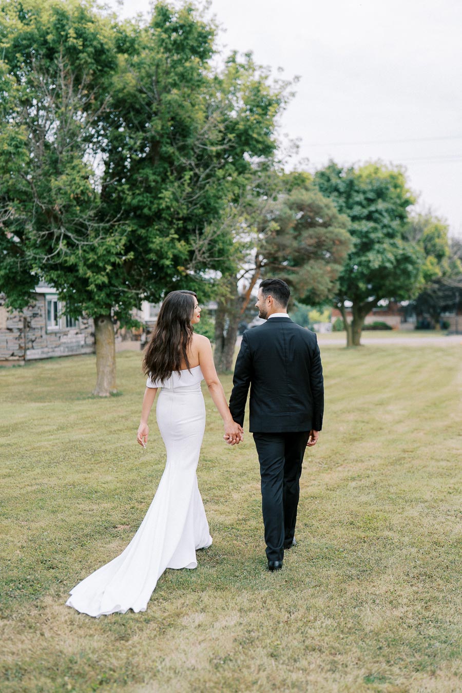 A couple walking hand in hand on a grassy field, with the woman in a white wedding dress and the man in a black suit, surrounded by green trees.