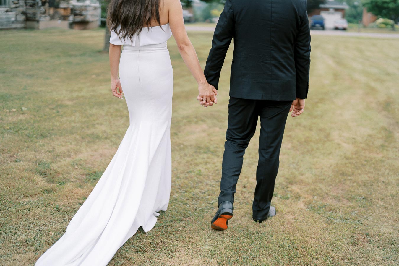 A couple holding hands walks on a grassy field; the woman is in a white wedding dress with a long train and the man is in a black suit.