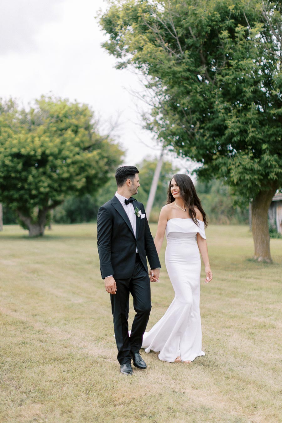 A bride and groom walking hand in hand on a grassy field, surrounded by green trees, with the bride wearing a white gown and the groom in a black suit.