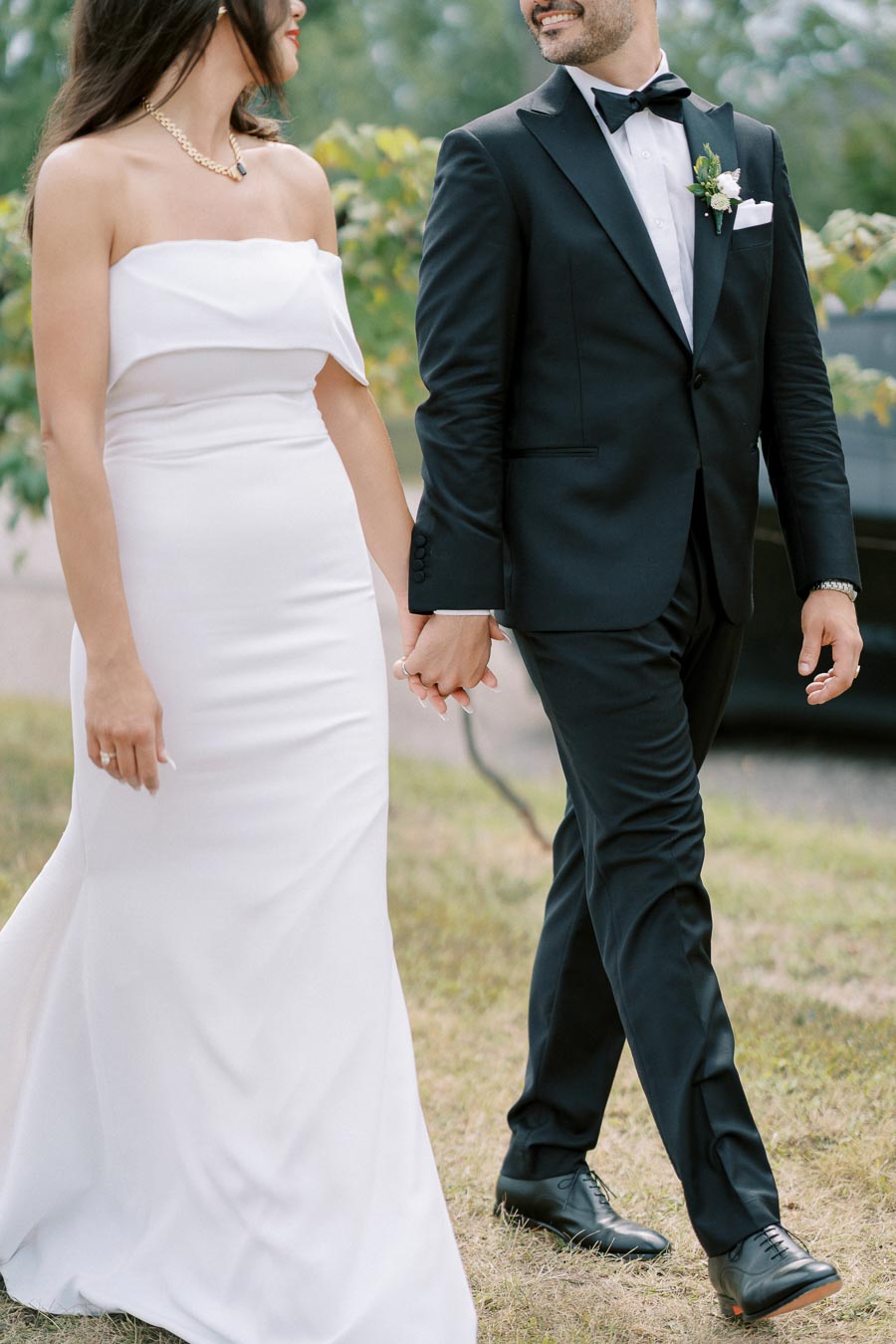 A couple in elegant wedding attire holding hands, with the bride in a strapless white dress and the groom in a black tuxedo, walking outdoors on a grassy path.