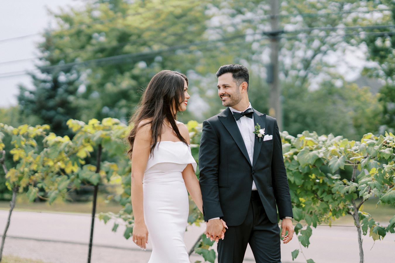 A bride in a white dress and groom in a black tuxedo smile at each other while holding hands amidst lush greenery outdoors, capturing a romantic wedding moment.