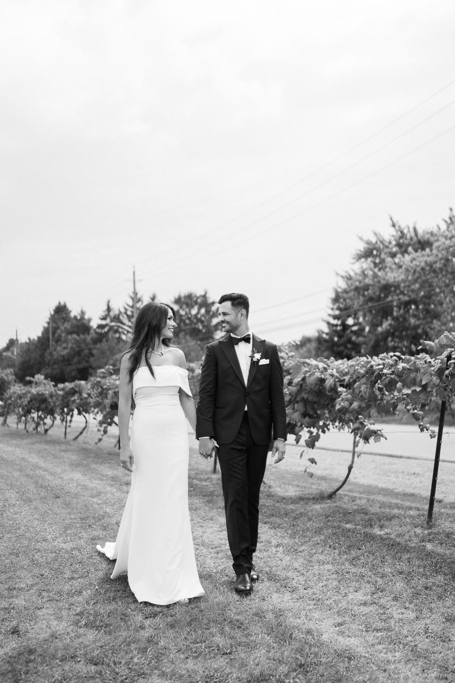 Black and white photo of a bride and groom holding hands and walking through a vineyard, both smiling at each other, with lush green vines in the background.