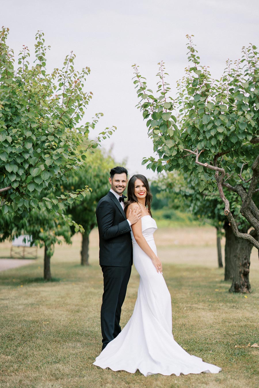 A couple in formal attire standing in a lush garden, with the man in a black suit and the woman in a white dress, smiling and embracing under green trees on a sunny day.