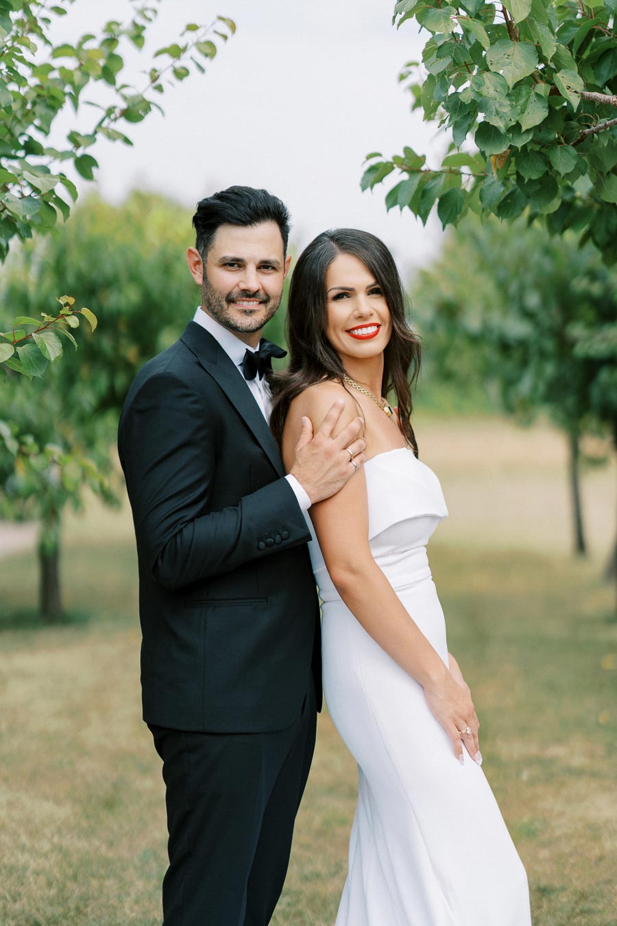 A couple posing outdoors, with the man wearing a black suit and bow tie, and the woman wearing a white dress, surrounded by green trees.