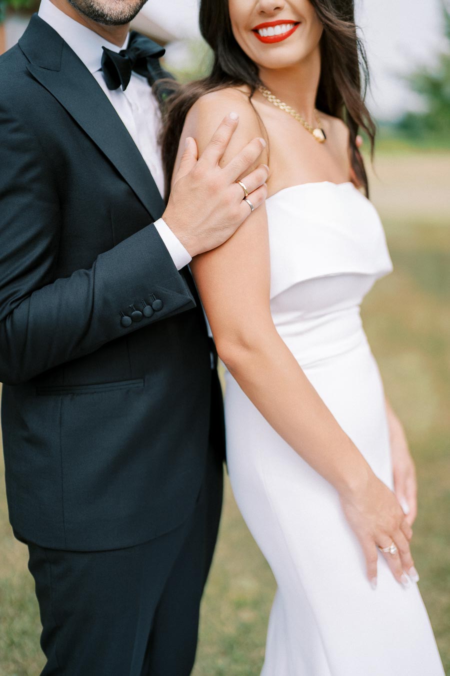 Bride and groom embracing, showcasing elegant wedding attire with the groom in a classic black tuxedo and the bride in a strapless white gown, highlighting rings and jewelry.