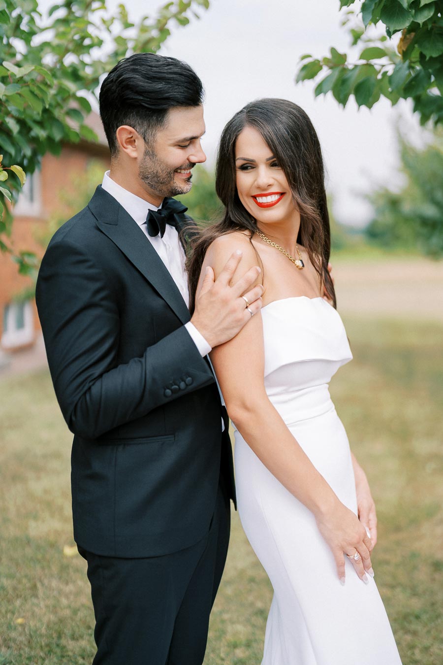 A smiling bride in a white gown stands with her groom in a black tuxedo, surrounded by greenery on their wedding day.