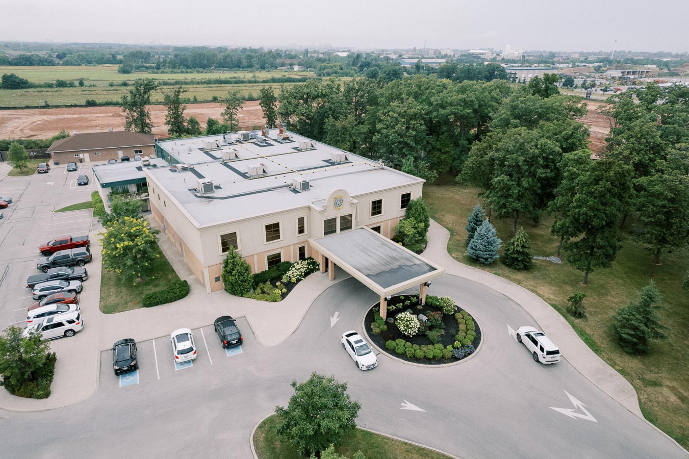 Aerial view of a modern commercial building with landscaped surroundings, parking area, and driveway leading to the main entrance, set amidst a suburban environment with green fields and trees in the background.