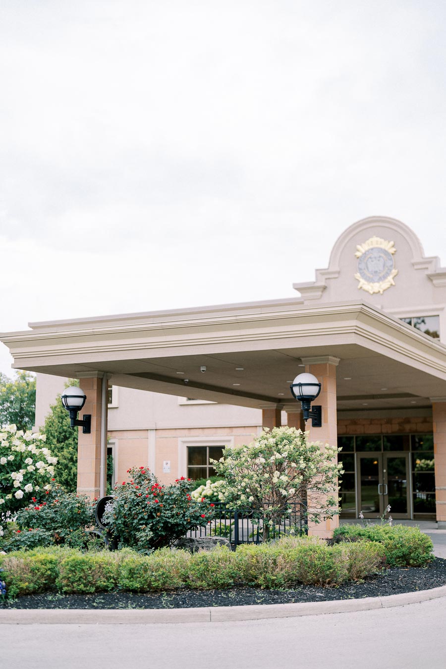 Elegant hotel entrance with manicured gardens and flowering bushes under a cloudy sky.
