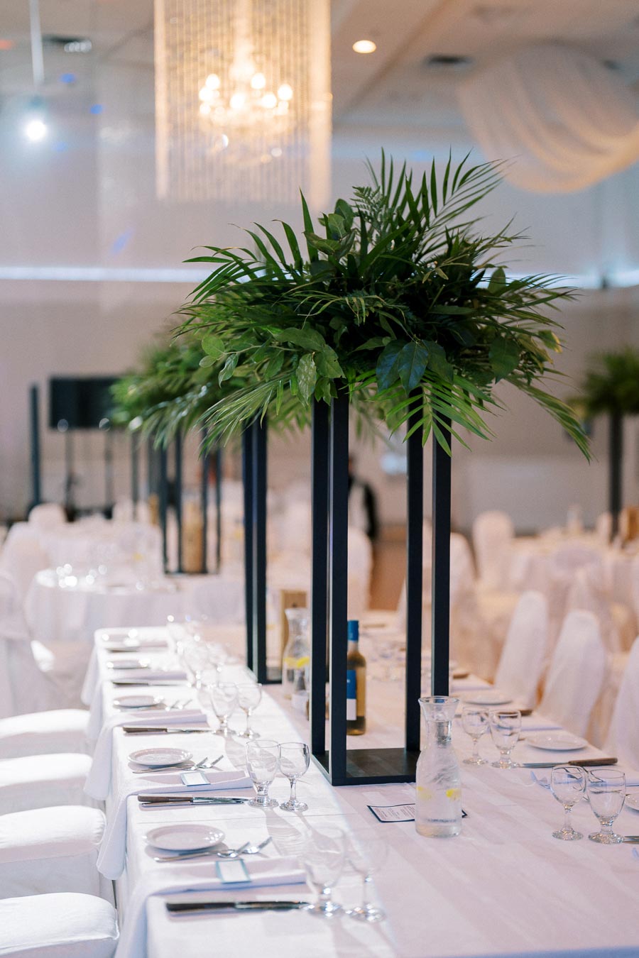 Elegant banquet table setting with white linens, adorned with tall centerpiece arrangements of lush green foliage, under a sparkling chandelier in a luxurious event space.