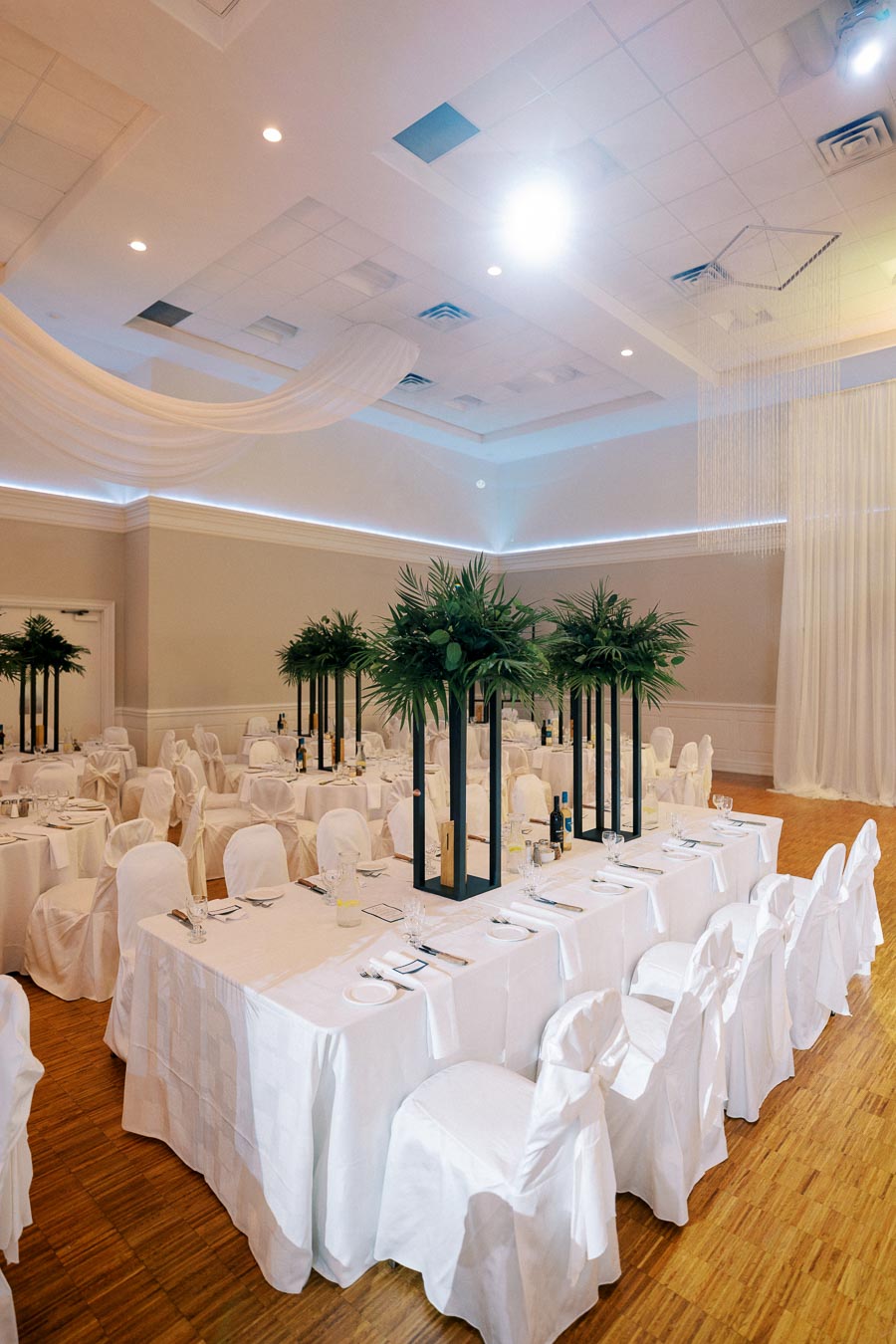 Elegant wedding reception hall with draped white linens and tall centerpieces featuring lush greenery, set in a spacious venue with a wooden floor and ambient lighting.