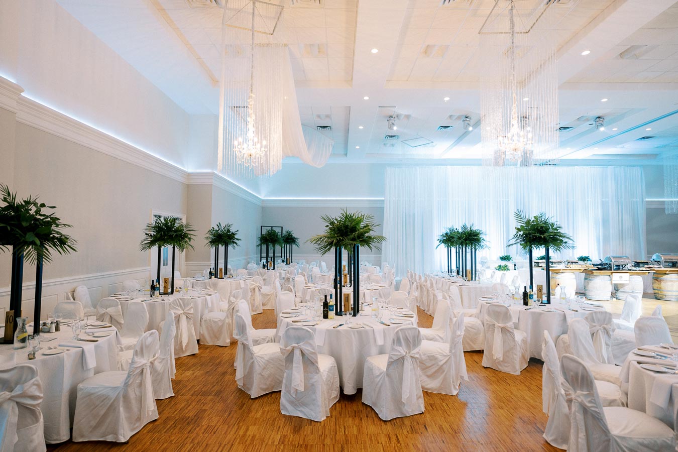 Elegant wedding reception venue with round tables and white chairs, adorned with tall floral centerpieces and chandeliers, set in a spacious, well-lit banquet hall with wooden floors and draped walls.