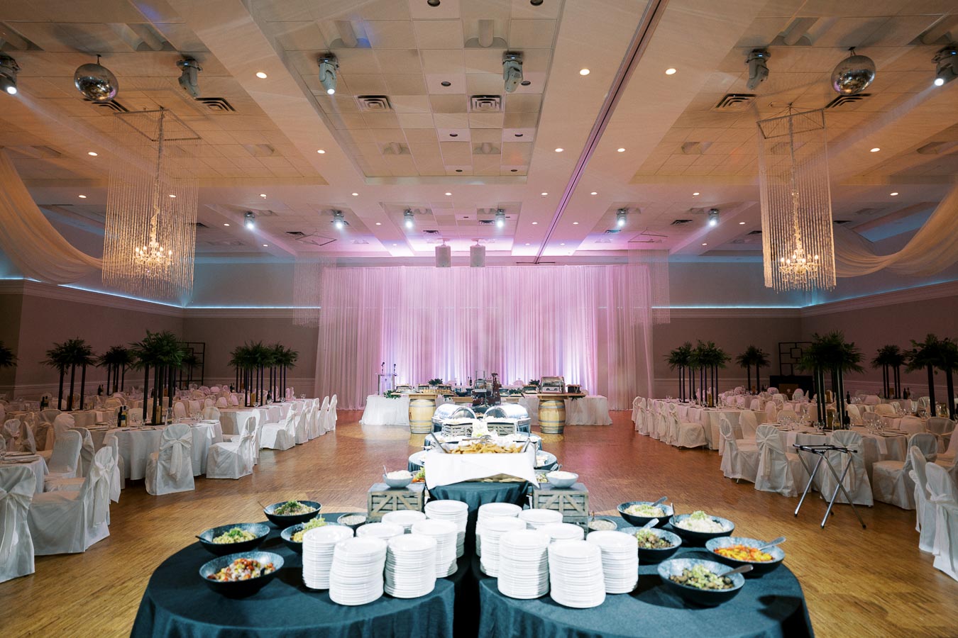 Elegant wedding reception venue with buffet setup, featuring round tables draped in white linens, decorated with tall greenery centerpieces. Soft, ambient lighting enhances the space with a pink backdrop and chandeliers creating a romantic atmosphere.