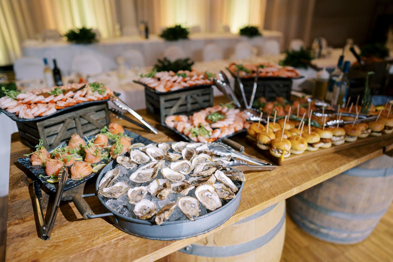 Elegant seafood and appetizer buffet with oysters, shrimp, smoked salmon rolls, and mini burgers at a wedding reception.