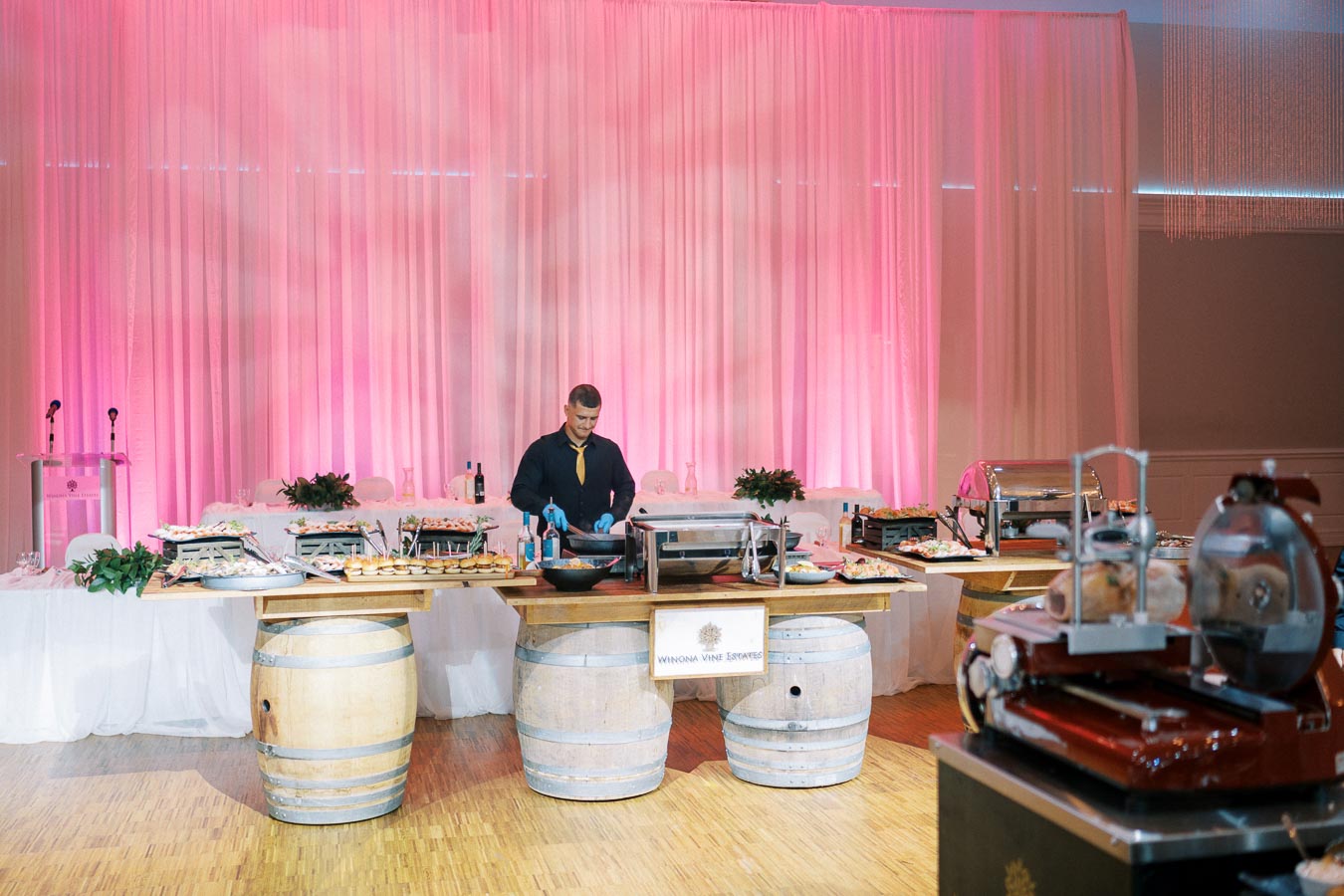 Elegant catering setup at an upscale event, featuring a chef preparing dishes behind a display of gourmet food platters and wine bottles, with ambient pink lighting and decorative wooden barrels in a spacious venue.