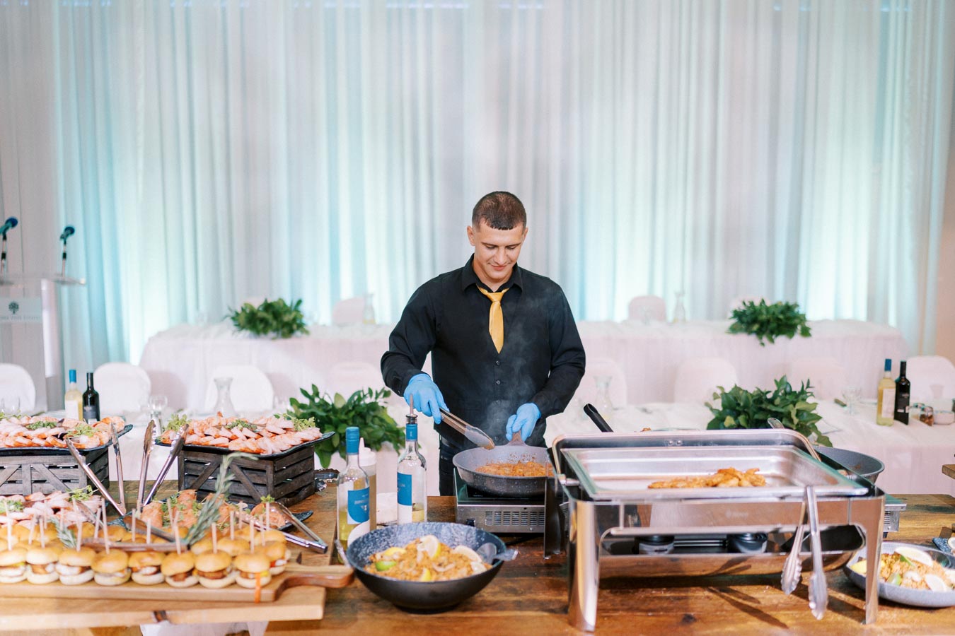 Chef preparing food at a catered event with various dishes and bottles on the table, wearing a black shirt with a yellow tie and blue gloves.