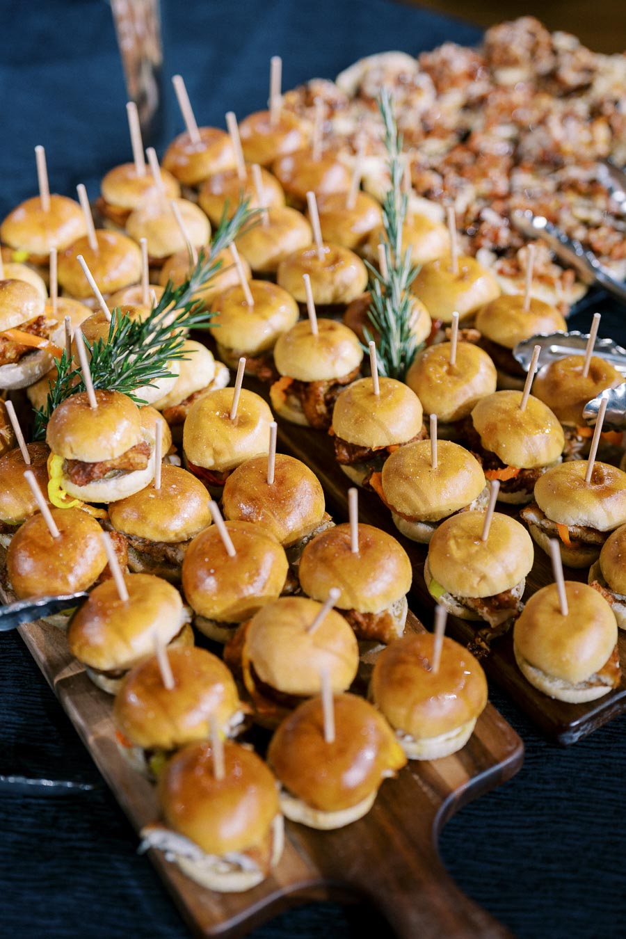 Platter of gourmet mini sliders topped with toothpicks, garnished with fresh rosemary sprigs on wooden serving boards, perfect for event catering or gourmet gatherings.