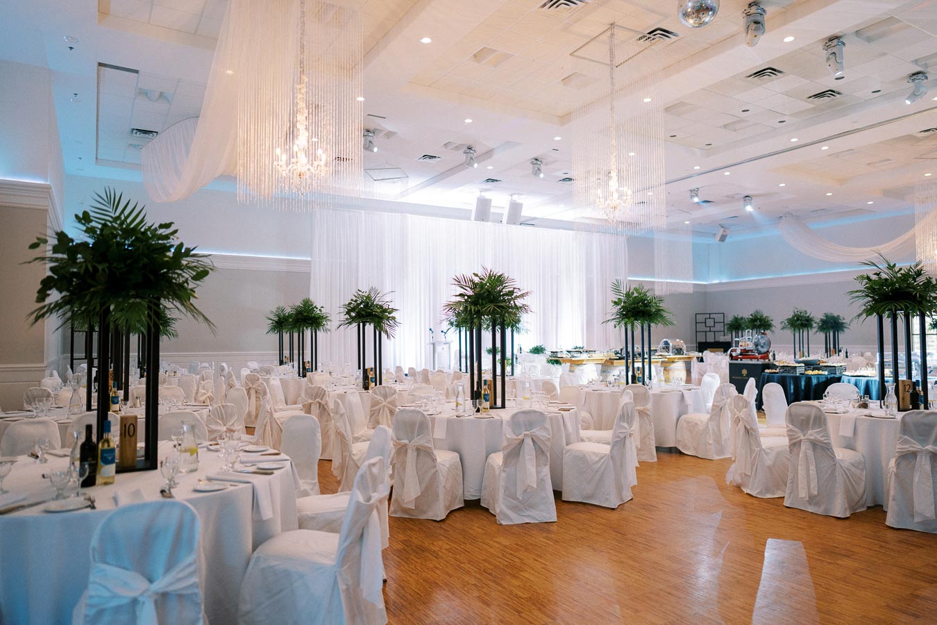 Elegant wedding reception hall with round tables draped in white linens and tall green centerpieces, featuring ambient lighting and a polished wooden floor, ideal for luxury events and celebrations.