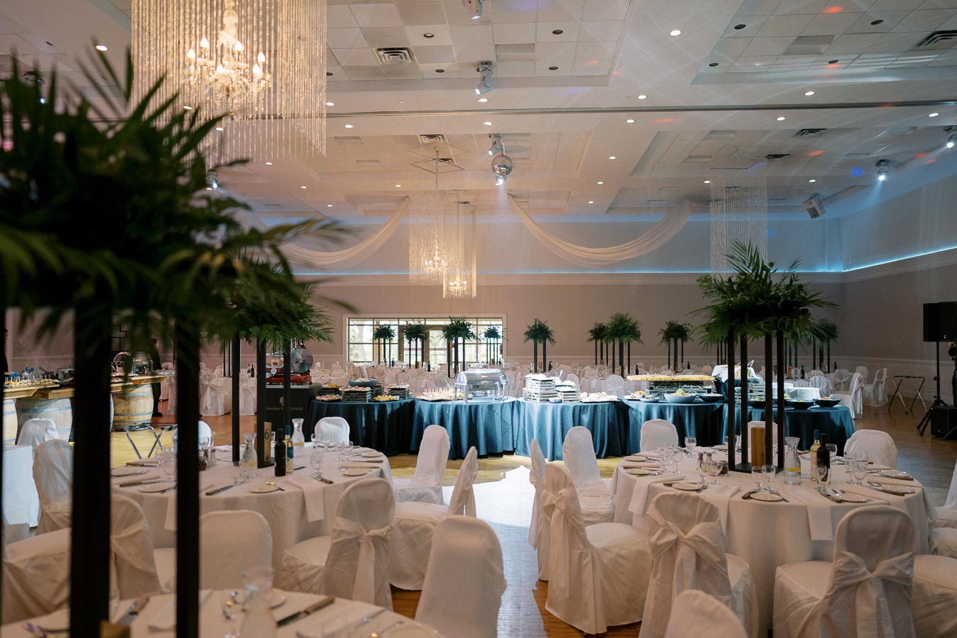 Luxurious event venue decorated with elegant white tablecloths, greenery centerpieces, and sparkling chandeliers, featuring round tables set for a formal dinner.
