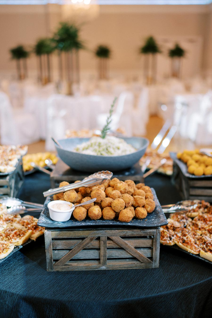 Elegant banquet setting with a variety of gourmet appetizers, including crispy fried balls, served on rustic wooden crates and a dark tablecloth, with decorative greenery in the background.