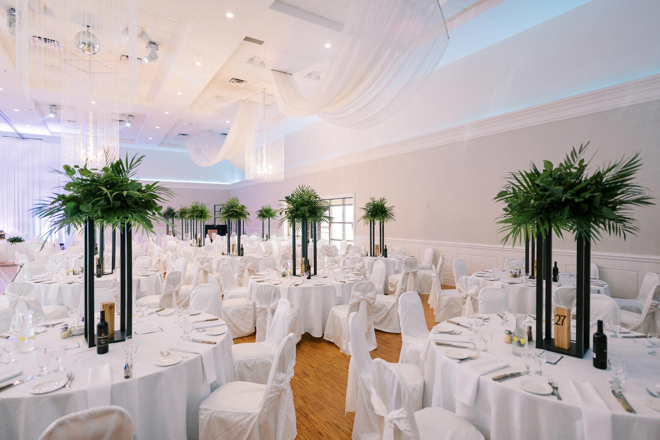 Elegant wedding reception hall with round tables adorned with white tablecloths and tall green centerpieces, under soft, flowing drapery and ambient lighting.